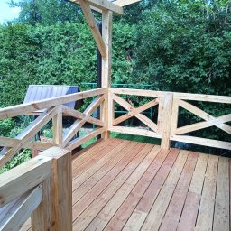 Tec-Dom - Nowo wybudowany taras z drewnianą podłogą i balustradą z krzyżowymi wzorami, otoczony zielenią.