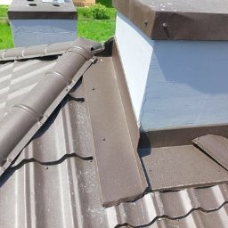 Tec-Dom - Brązowe orynnowanie i blaszane obróbki komina na dachu z metalowej dachówki w słoneczny dzień.