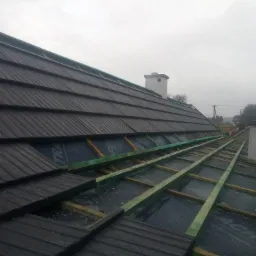 Fragment dachu w trakcie montażu, widoczne czarne dachówki, zielone łaty, czarna membrana dachowa oraz komin w tle, pochmurne niebo.