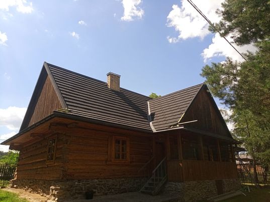 Drewniany dom z ciemnobrązowym dachem, widok z boku w słoneczny dzień, z widocznym kominem i fragmentem werandy.