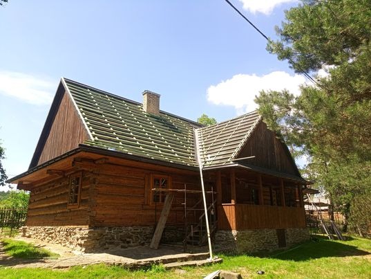 Drewniany dom z widoczną konstrukcją dachu przygotowaną do położenia nowego pokrycia, komin murowany, podmurówka z kamienia, rusztowania przy ścianie.