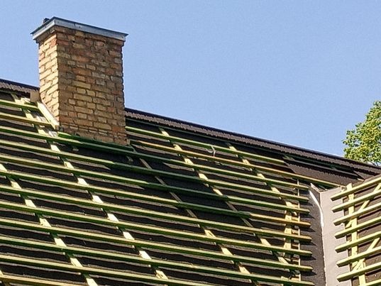 Konstrukcja więźby dachowej z kominem z cegły w trakcie montażu, widoczne łaty i kontrłaty na tle błękitnego nieba.