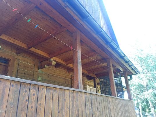 Drewniana weranda domu z widocznymi detalami konstrukcyjnymi, czarną rynną i sznurkiem z kolorowymi klamerkami.