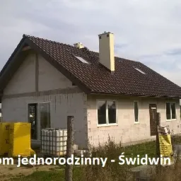 Surowy stan domu jednorodzinnego z brązowym dachem, widoczne palety z materiałami budowlanymi i zbiornik na wodę, w tle polny krajobraz.