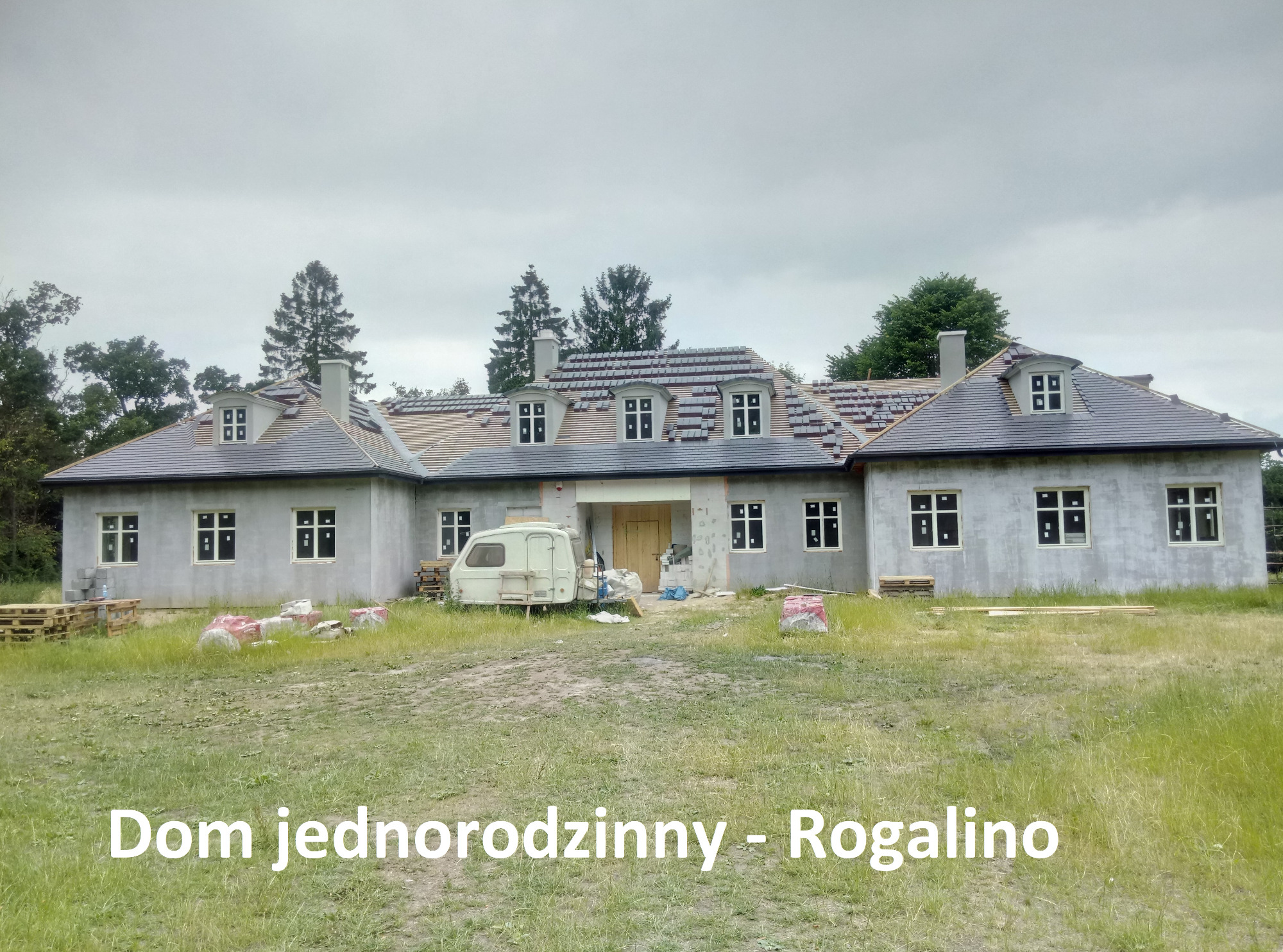 Surowy stan domu jednorodzinnego z szarą elewacją i częściowo ułożonym dachem z dachówki, widoczny przyczepka i materiały budowlane na trawniku.