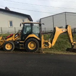 Żółta koparko-ładowarka CAT 428F2 zaparkowana na poboczu asfaltowej drogi, z widocznymi numerami 88852 i 8567 na kabinie, w tle dom jednorodzinny i hala.