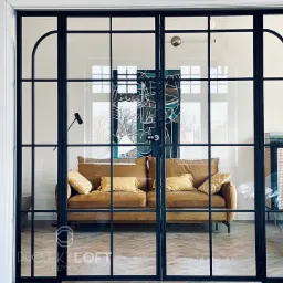 Dwuskrzydłowe drzwi w stylu loftowym z czarnymi, metalowymi szprosami i przeszkleniami, widok na salon z brązową sofą i parkietem w jodełkę, logo firmy w dolnym rogu.