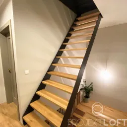 Nowoczesne schody ażurowe z drewnianymi stopniami i czarną metalową konstrukcją boczną, widoczne z dołu, w tle ściana i drzwi.