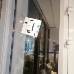 Biały robot myjący okna, model Hobot 298, przyczepiony do szyby okiennej, z widocznym kablem zasilającym i delikatnymi smugami czyszczącego płynu.