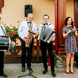 Czteroosobowy zespół muzyczny w eleganckich strojach grający na zewnątrz budynku z czerwonym dywanem, saksofonista z mikrofonem na statywie, akordeonista, perkusista z pałką i wokalistka z tamburynem.