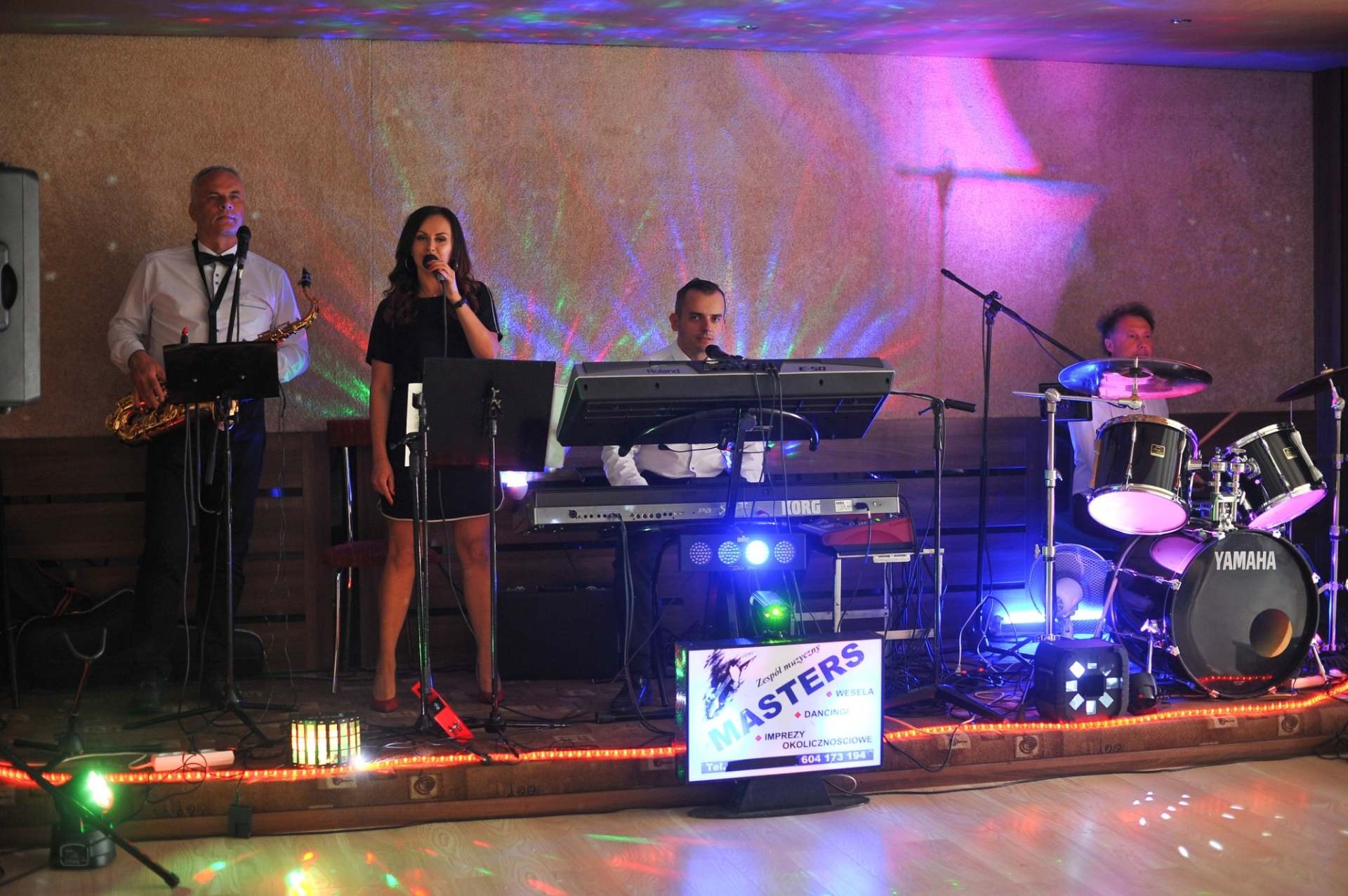 Zespół muzyczny na scenie podczas występu: wokalistka śpiewa do mikrofonu, saksofonista gra na saksofonie, klawiszowiec gra na keyboardzie, a perkusista na perkusji Yamaha. Scena oświetlona...