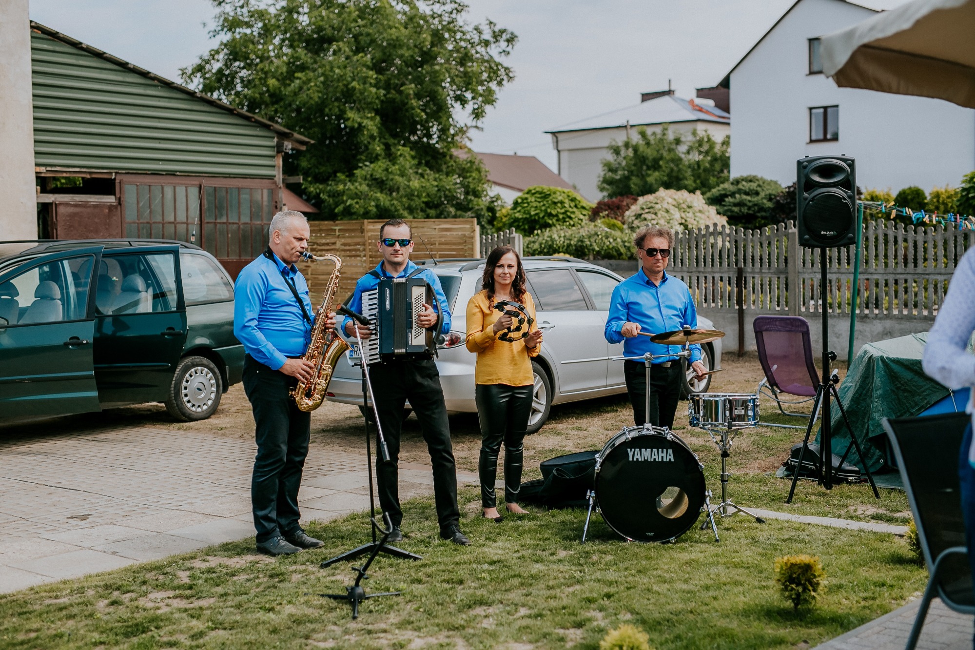 Czteroosobowy zespół muzyczny grający na zewnątrz: saksofonista, akordeonista, tamburynistka i perkusista z talerzami, z samochodami i budynkami w tle.
