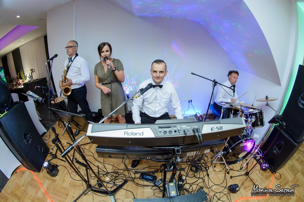 Zespół muzyczny w trakcie występu: saksofonista, wokalistka, klawiszowiec grający na Roland E-50 i perkusista, oświetlenie sceniczne, widoczne kable i sprzęt nagłośnieniowy.