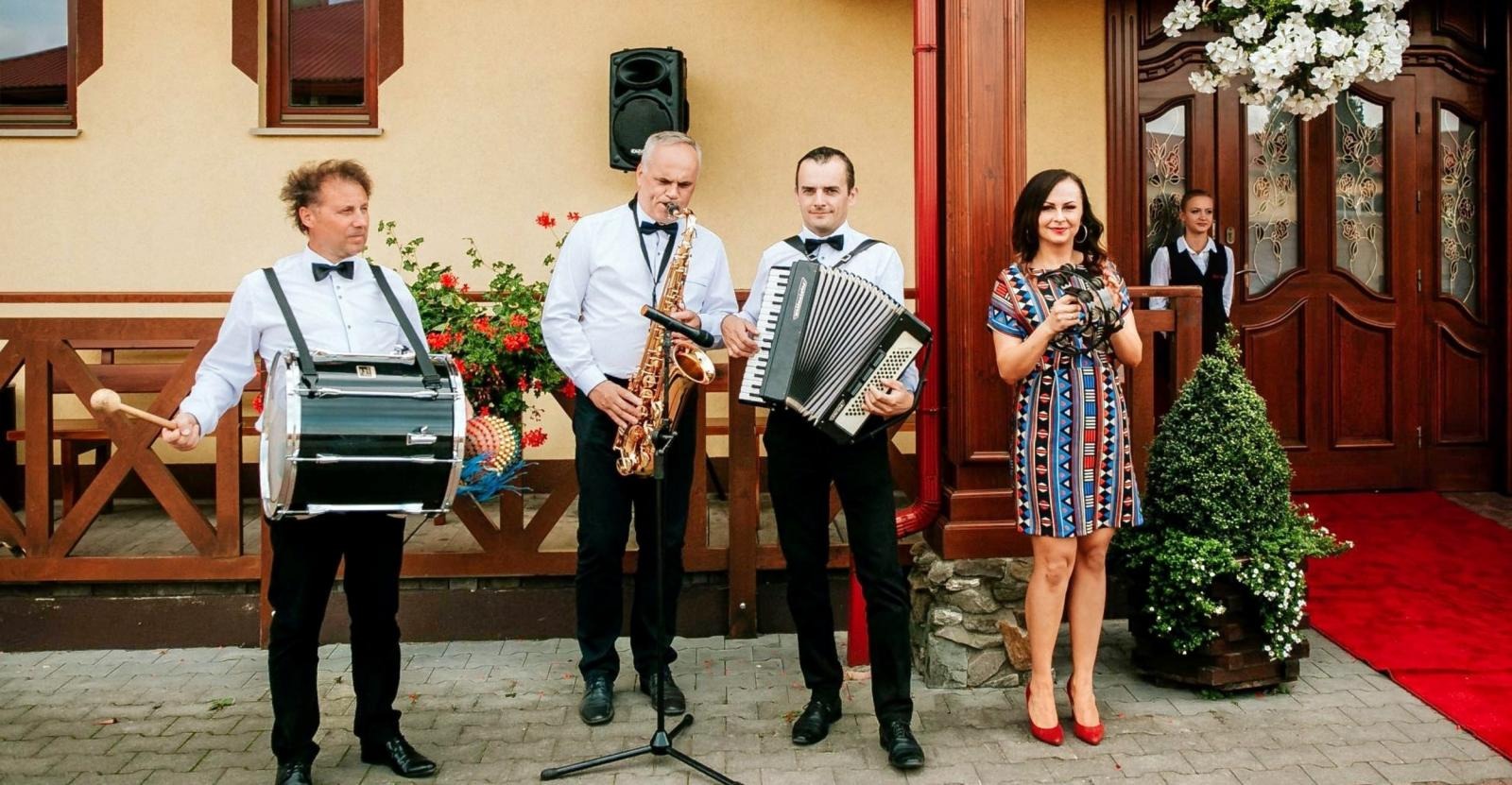 Czteroosobowy zespół muzyczny w eleganckich strojach grający na zewnątrz budynku z czerwonym dywanem, saksofonista z mikrofonem na statywie, akordeonista, perkusista z pałką i wokalistka z tamburynem.