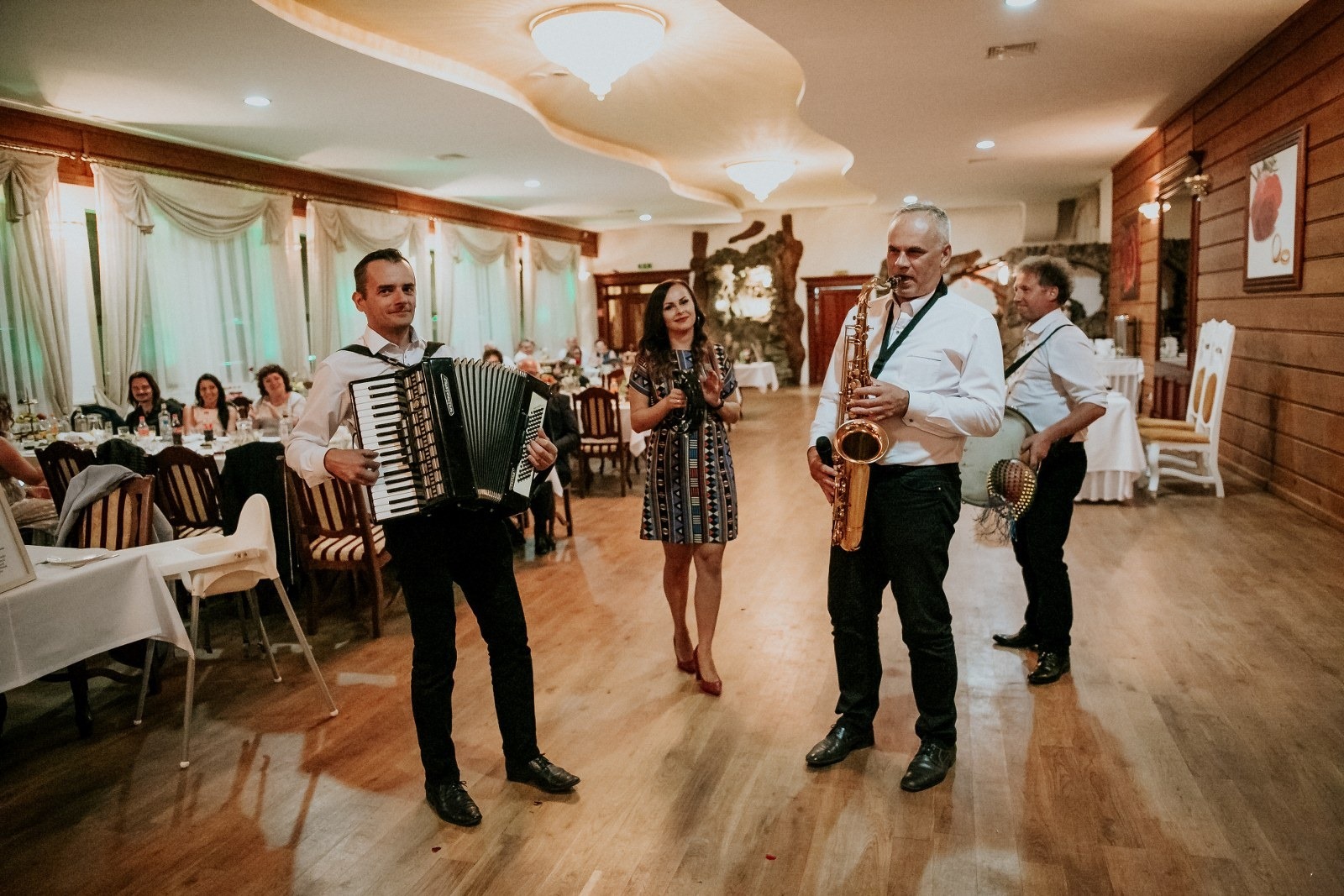 Zespół muzyczny grający na weselu: akordeonista, saksofonista i wokalistka na parkiecie, w tle goście siedzący przy stołach.