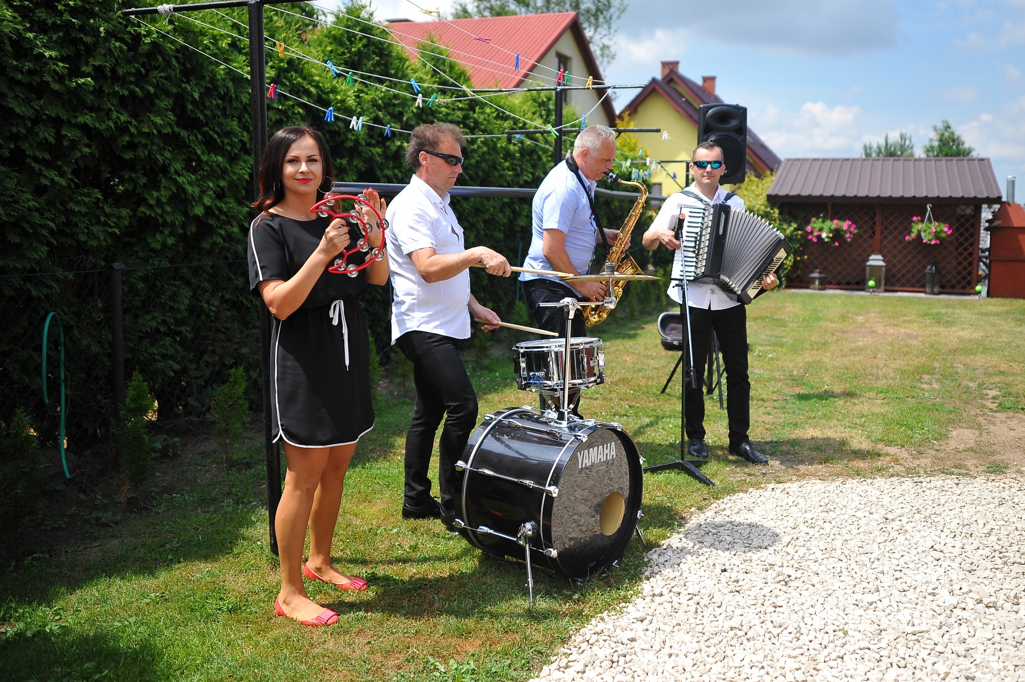 Czteroosobowy zespół grający na trawniku: kobieta z tamburynem, perkusista, saksofonista i akordeonista, za nimi żywopłot i dom.