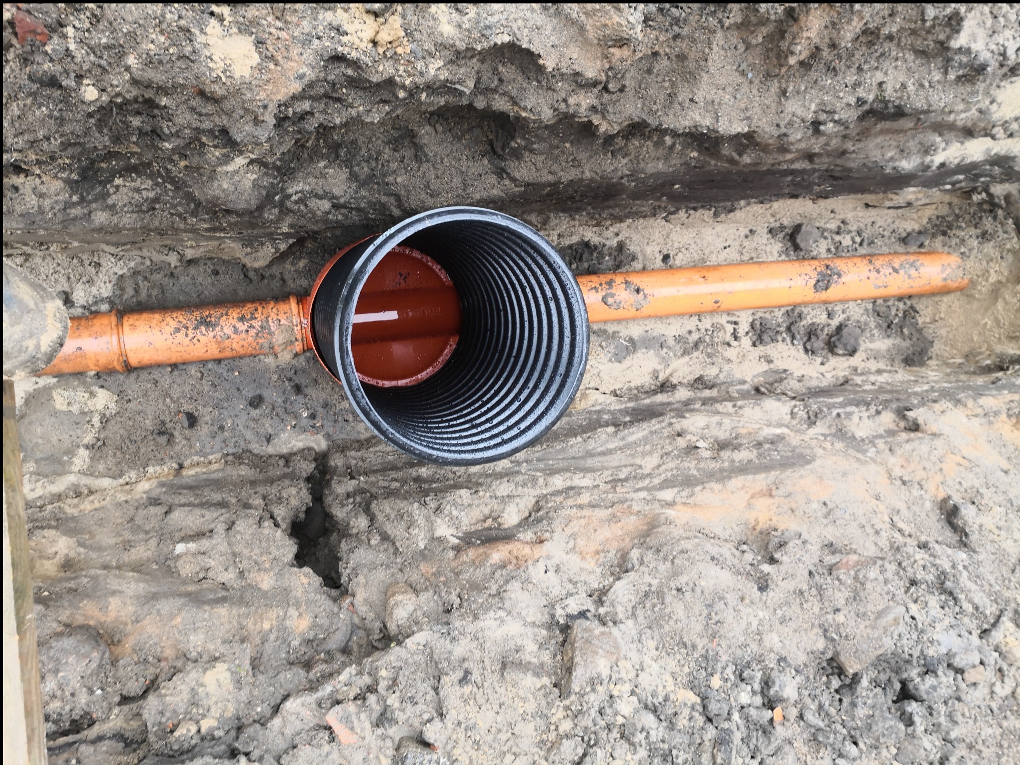 Pomarańczowa rura kanalizacyjna z czarnym karbowanym zakończeniem, ułożona w wykopie ziemnym. Widoczne warstwy gleby i fragmenty gruzu.