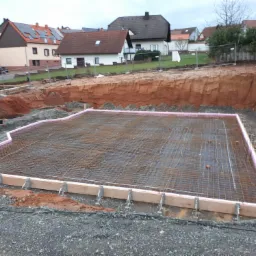 Przygotowanie fundamentów pod budynek: wykop, siatka zbrojeniowa, obrzeża z izolacją różową i szalunkiem drewnianym, grunt wokół wykopu.