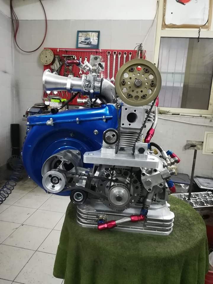 Złożony, odrestaurowany silnik samochodowy z niebieską obudową i srebrnymi elementami, widoczny na stole warsztatowym z zielonym obrusem, na tle ściany z narzędziami i zegarem.