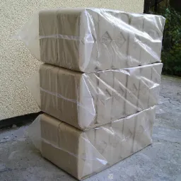 Trzy ułożone jeden na drugim pakiety brykietu dębowego RUF, zabezpieczone przezroczystą folią, na betonowym podłożu, z fragmentem ściany budynku w tle.