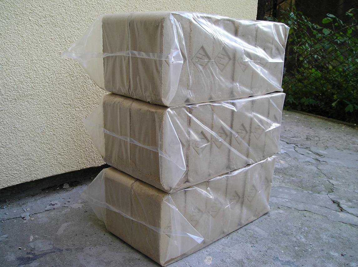 Trzy ułożone jeden na drugim pakiety brykietu dębowego RUF, zabezpieczone przezroczystą folią, na betonowym podłożu, z fragmentem ściany budynku w tle.