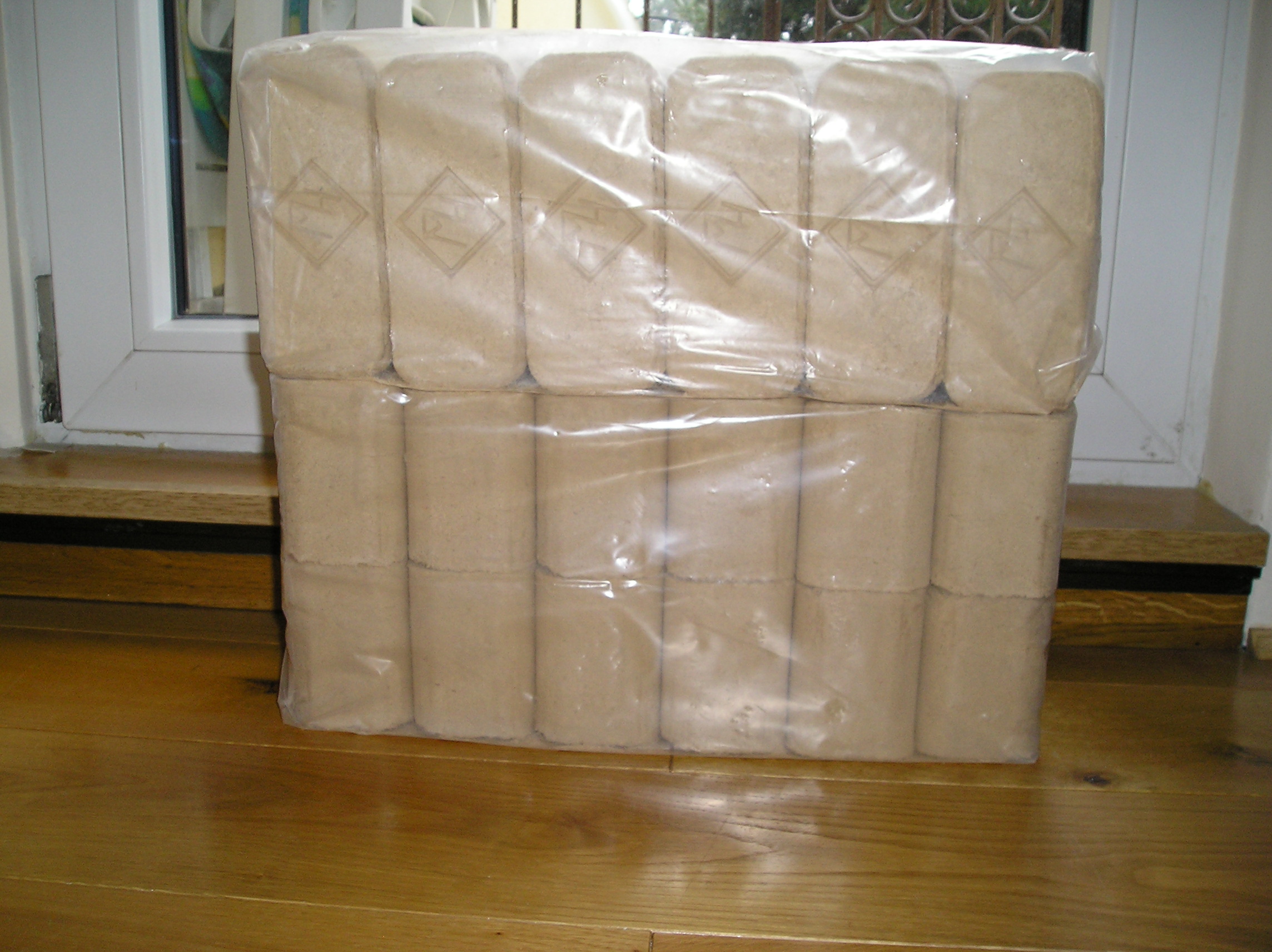 Pakiet brykietu dębowego RUF owinięty folią, ułożony na drewnianej podłodze w pobliżu okna.