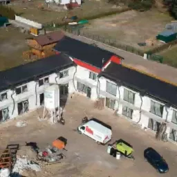 Widok z drona na budynek w trakcie ocieplania, z widocznym agregatem do pianki PUR, samochodem dostawczym i pracownikami na placu budowy.