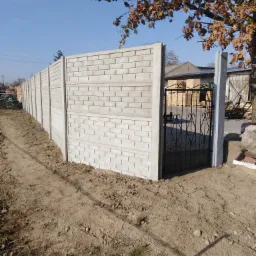 Długi, prefabrykowany, betonowy płot z wzorem cegły i czarną, metalową furtką wejściową, widziany z zewnątrz posesji na tle jesiennego krajobrazu.