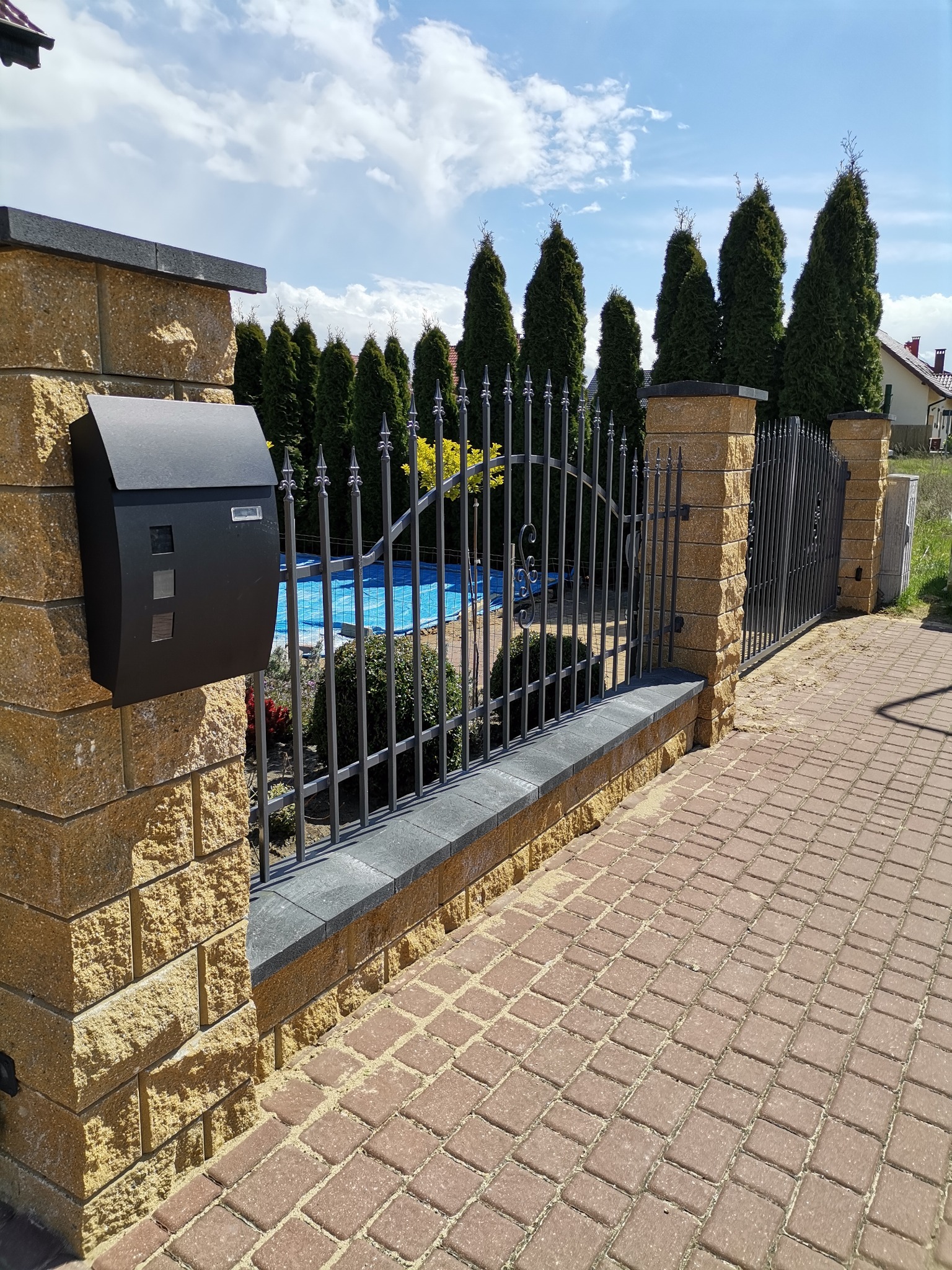 Nowoczesne ogrodzenie z elementami kutymi, słupki z kostki łupanej w kolorze piaskowym, skrzynka pocztowa i chodnik z kostki brukowej w Kaczorach.