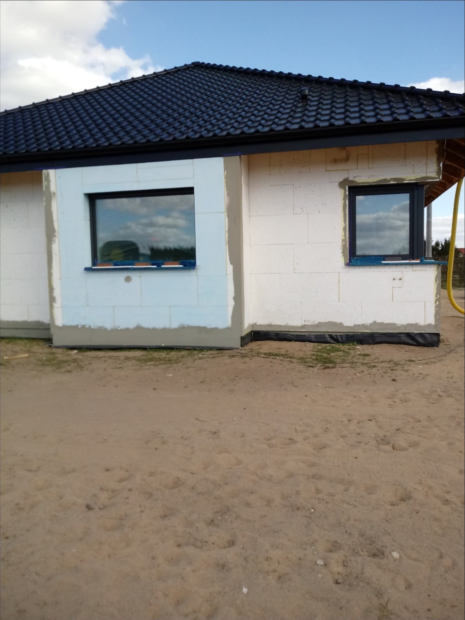 Fragment elewacji domu w trakcie ocieplania, widoczne białe bloczki, częściowo otynkowane i ocieplone, z zamontowanymi oknami z ciemnymi ramami.