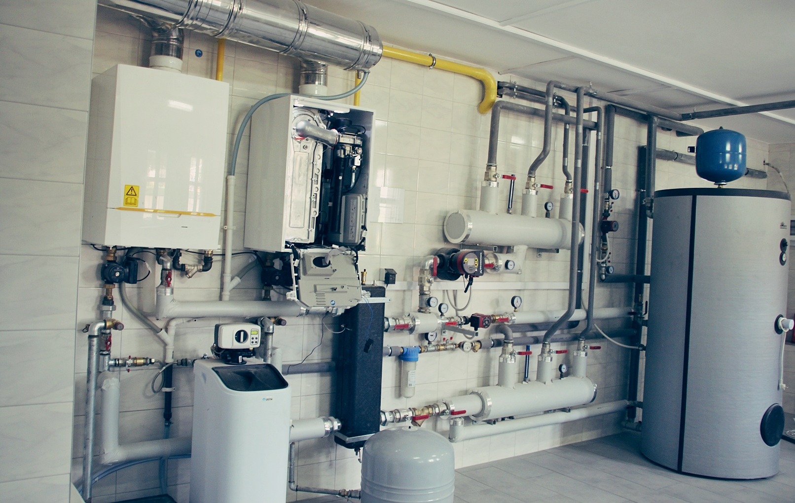 Kompleksowa instalacja grzewcza z dwoma piecami gazowymi, zbiornikiem na wodę i licznymi rurami oraz manometrami na kafelkowanej ścianie.