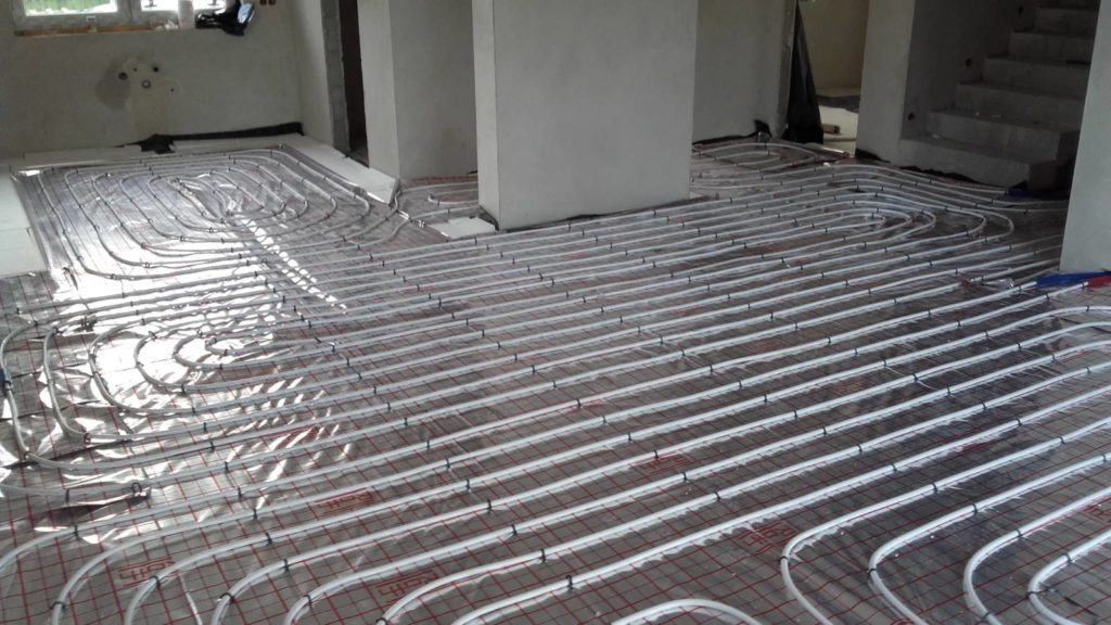 Rozłożona instalacja ogrzewania podłogowego w surowym wnętrzu, rury grzewcze ułożone równolegle na folii z siatką, widoczne fragmenty ścian i schodów w stanie deweloperskim.