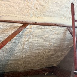 IZOLACJE PIANĄ PUR - Poddasze z widoczną izolacją pianką PUR, drewniane elementy konstrukcyjne, fragment muru z cegły. Aplikacja pianki w trudno dostępnym miejscu.