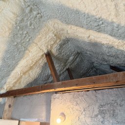 IZOLACJE PIANĄ PUR - Poddasze z drewnianą konstrukcją pokryte grubą warstwą jasnej pianki izolacyjnej, widoczne fragmenty ściany z lampą. Detal izolacji natryskowej.