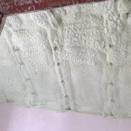 IZOLACJE PIANĄ PUR - Poddasze po ociepleniu pianką: gęsta, jasna warstwa izolacyjna pokrywa całą powierzchnię, widoczne fragmenty cegieł i ściany. Tekstura pianki przypomina gąbkę.