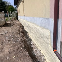 IZOLACJE PIANĄ PUR - Ocieplenie fundamentu pianką PUR na ścianie budynku z widoczną rurą spustową i wykopem wzdłuż ściany. Widoczne ogrodzenie i drzewa w tle.