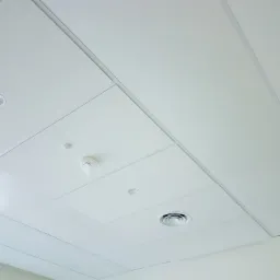 Kasetonowy sufit podwieszany w kolorze białym z widocznym czujnikiem dymu w folii ochronnej, okrągłą kratką wentylacyjną i punktowym oświetleniem.