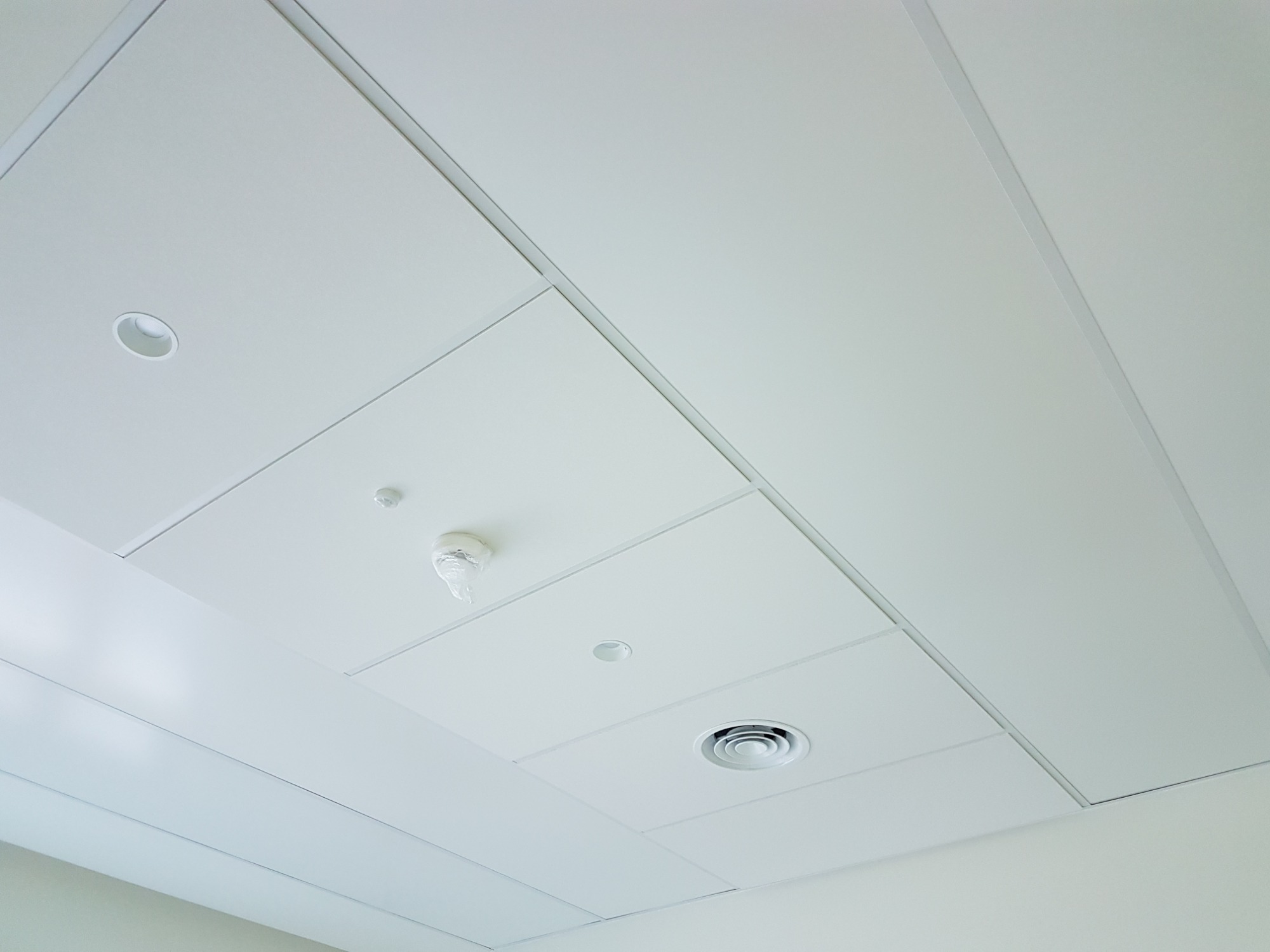 Kasetonowy sufit podwieszany w kolorze białym z widocznym czujnikiem dymu w folii ochronnej, okrągłą kratką wentylacyjną i punktowym oświetleniem.