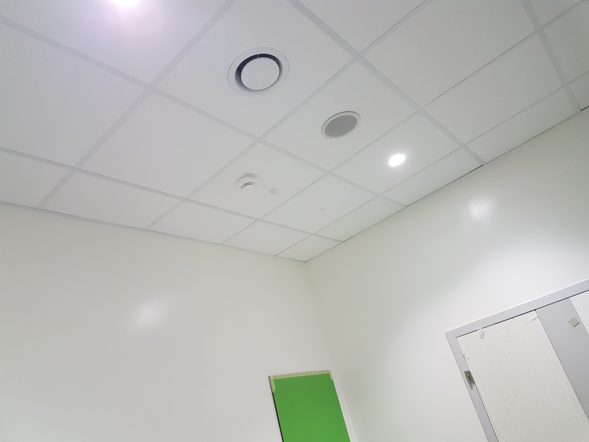 Białe pomieszczenie biurowe po odświeżeniu, widoczny sufit kasetonowy z zamontowanymi punktami oświetleniowymi i kratką wentylacyjną, ściany pomalowane na biało, fragment zabezpieczonej taśmą...
