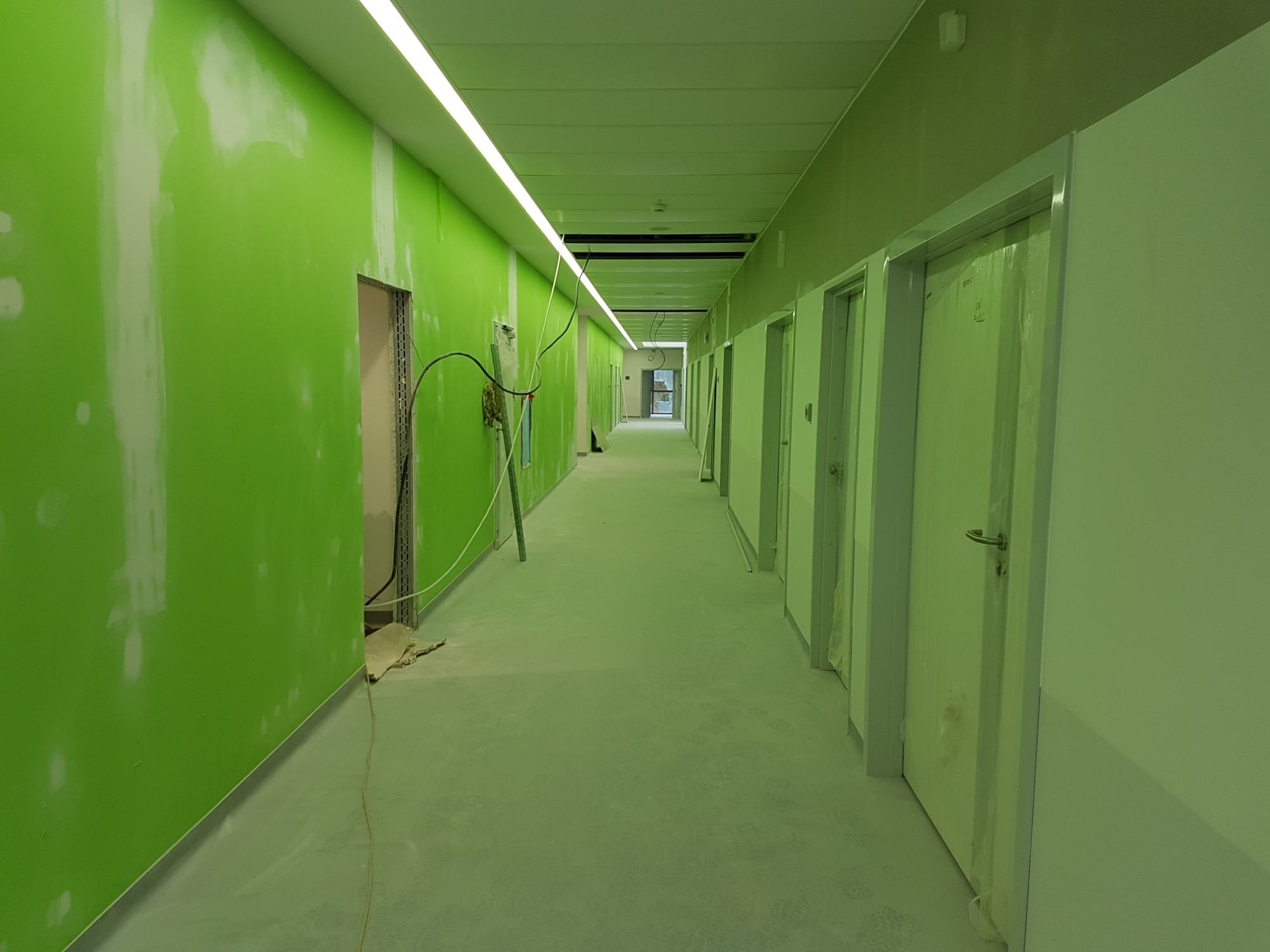 Korytarz w trakcie prac wykończeniowych, ściany pomalowane na jaskrawy zielony kolor z widocznymi śladami po szpachlowaniu, drzwi zabezpieczone folią, widoczne kable i przewody.