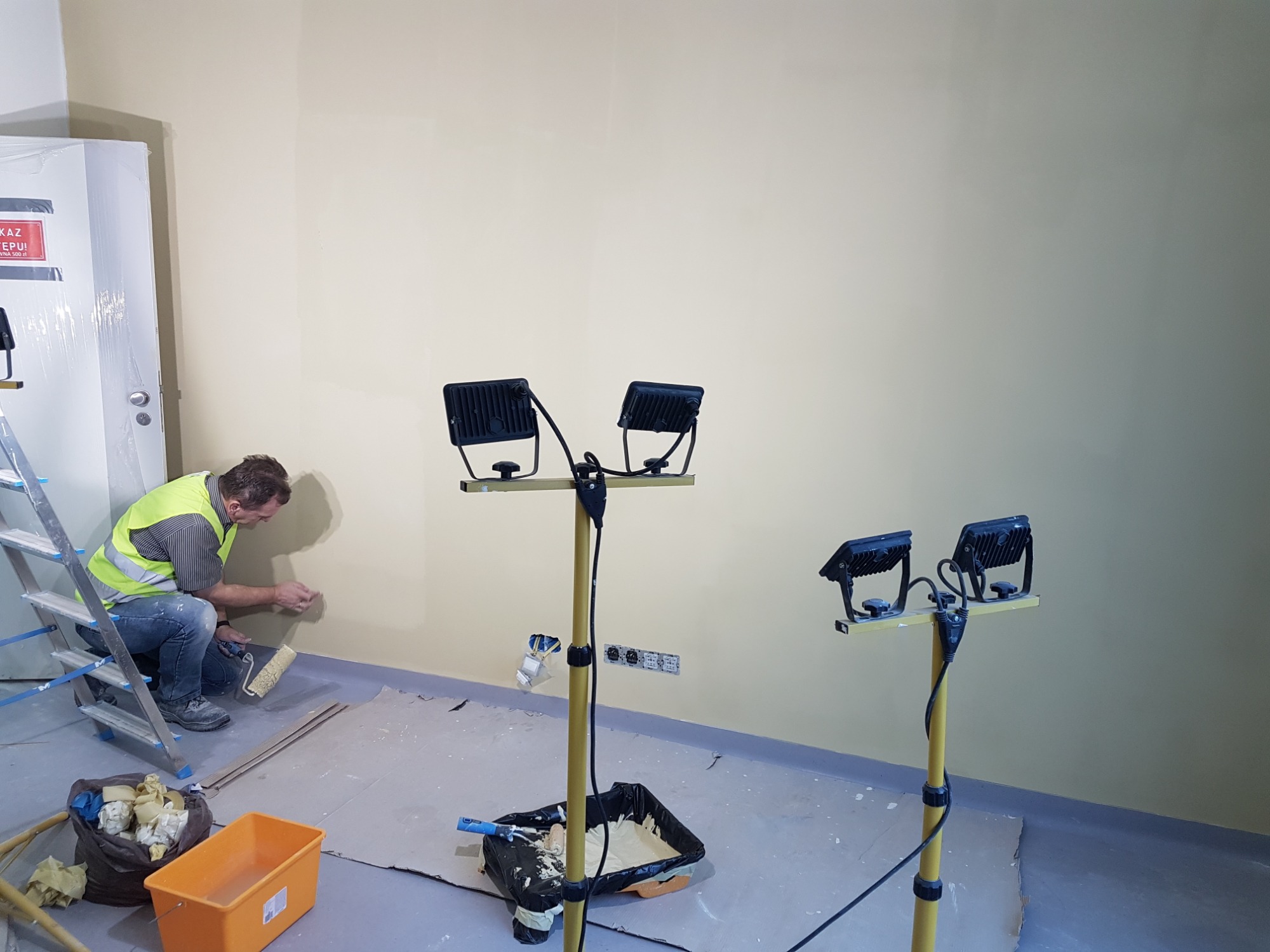 Mężczyzna w kamizelce odblaskowej, siedząc na drabinie, maluje ścianę wałkiem w pomieszczeniu oświetlonym lampami budowlanymi. Widoczny pojemnik z farbą i akcesoria malarskie.