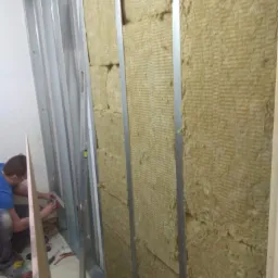 Montaż izolacji z wełny mineralnej w metalowym stelażu pod płytę karton-gips, widoczne narzędzia i poziomica.