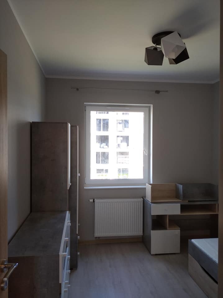 Pokój po odświeżeniu, szare ściany, nowoczesna lampa sufitowa z geometrycznymi kloszami, drewnopodobna podłoga, białe okno z widokiem na budynek, grzejnik pod oknem, szafa z szufladami i biurko...