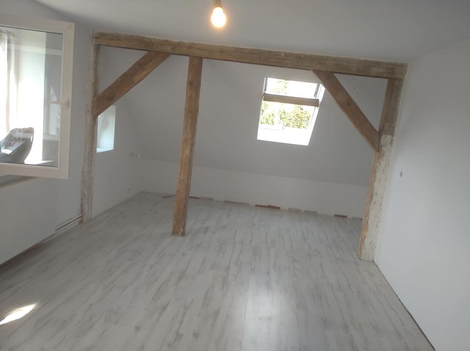 Poddasze po remoncie z drewnianą konstrukcją nośną, wykończone na biało, z nową podłogą imitującą drewno i oknem dachowym.