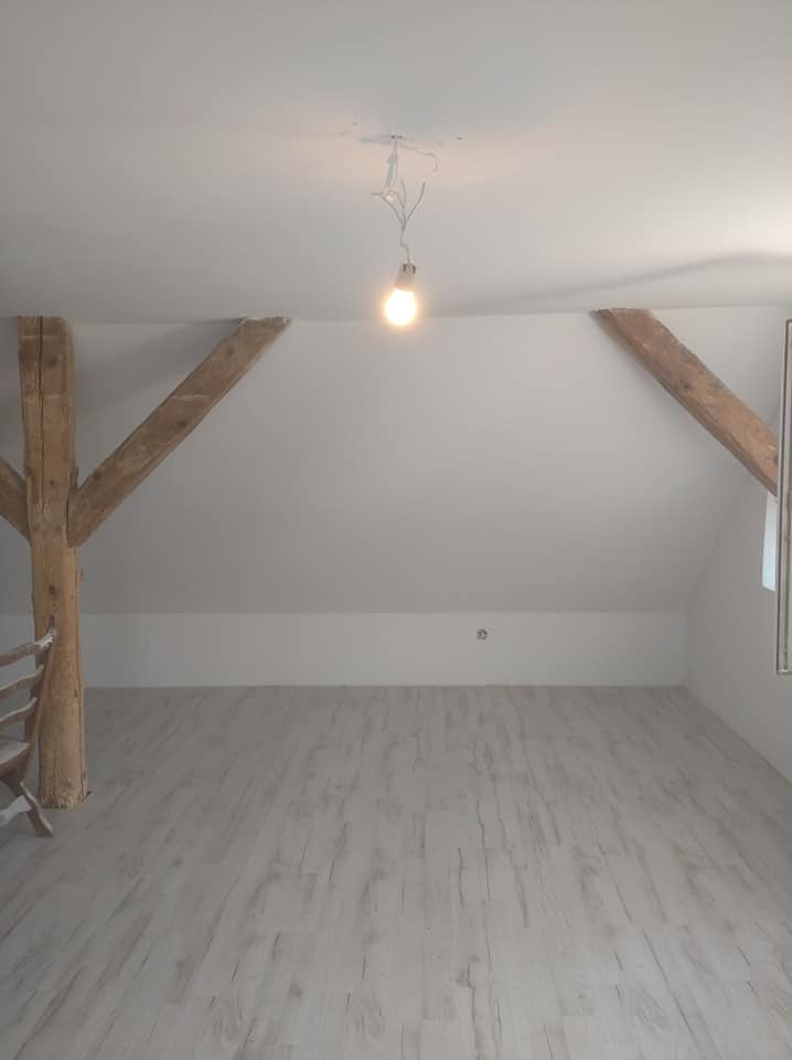 Poddasze po remoncie z drewnianymi belkami stropowymi, świeżo pomalowane na biało ściany i sufit, jasna podłoga imitująca drewno, wisząca żarówka na kablu.