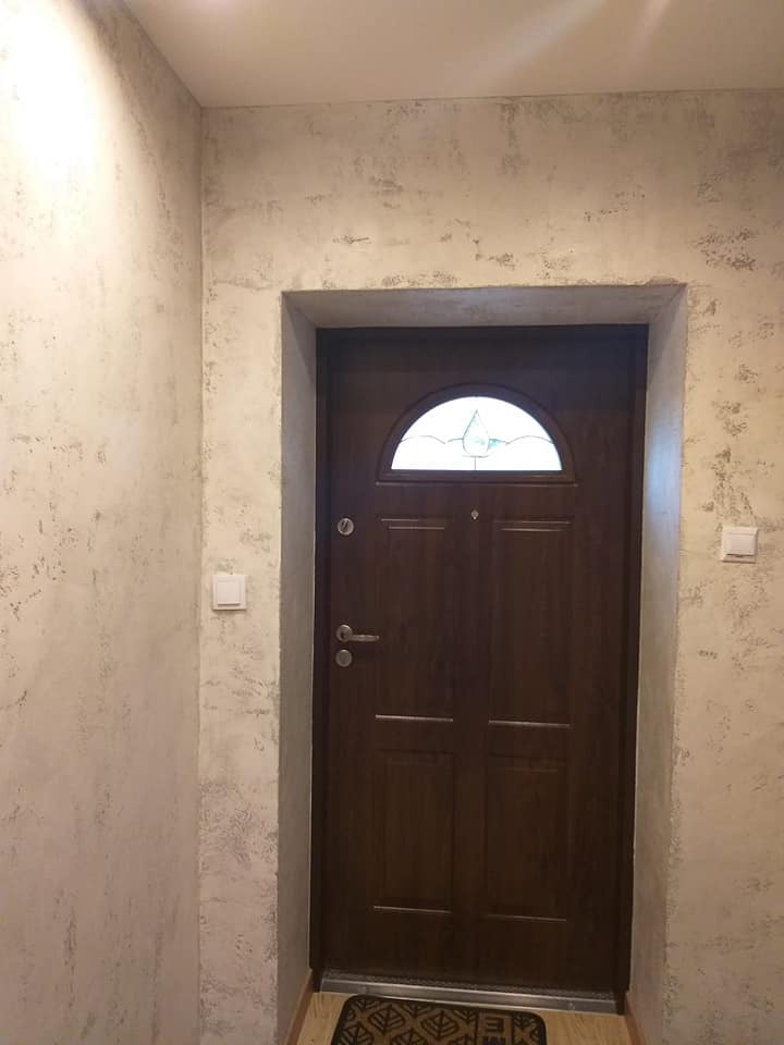 Ciemne, drewniane drzwi wejściowe z półokrągłym przeszkleniem, osadzone w otynkowanej ścianie o nieregularnej fakturze, widoczne w korytarzu z włącznikami światła po obu stronach.