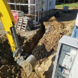 Żółta mini koparka wykonuje wąski wykop pod instalację elektryczną, obok otwarta skrzynka rozdzielcza z licznikiem Norax 3 i bezpiecznikami, w tle budynek w trakcie budowy z białego bloczka.