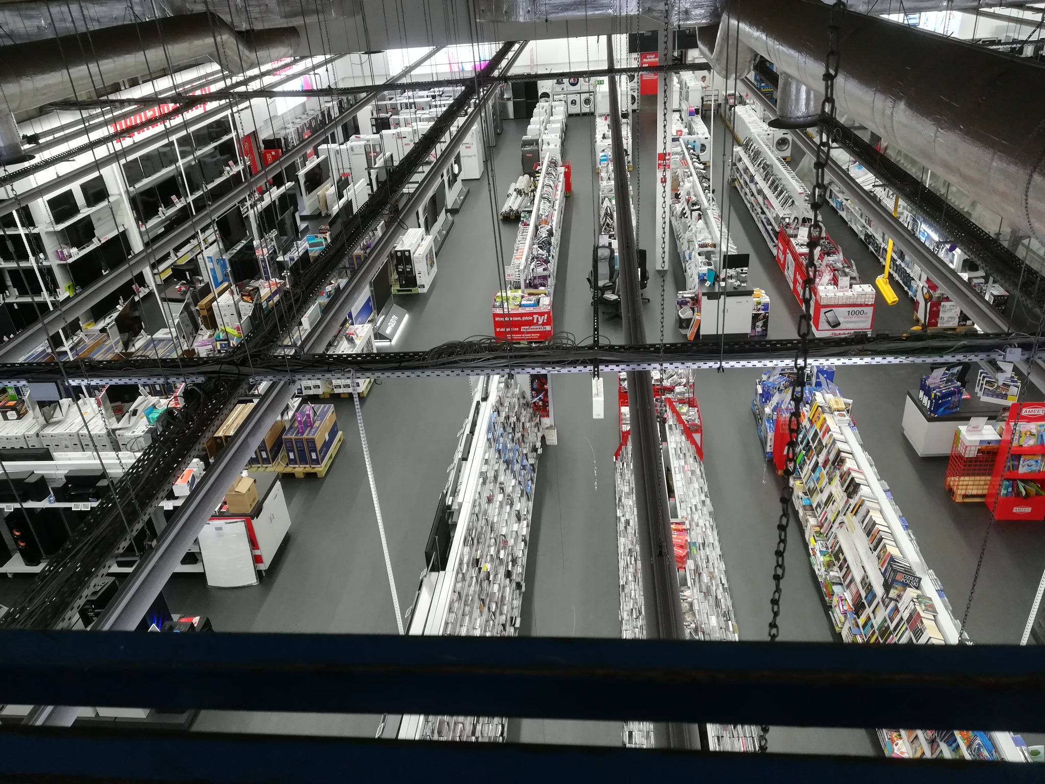 Widok z góry na wnętrze sklepu z elektroniką i sprzętem AGD, widoczne regały z telewizorami, pralkami, suszarkami i drobnym sprzętem, oświetlenie sufitowe, metalowe rury i łańcuchy pod sufitem.