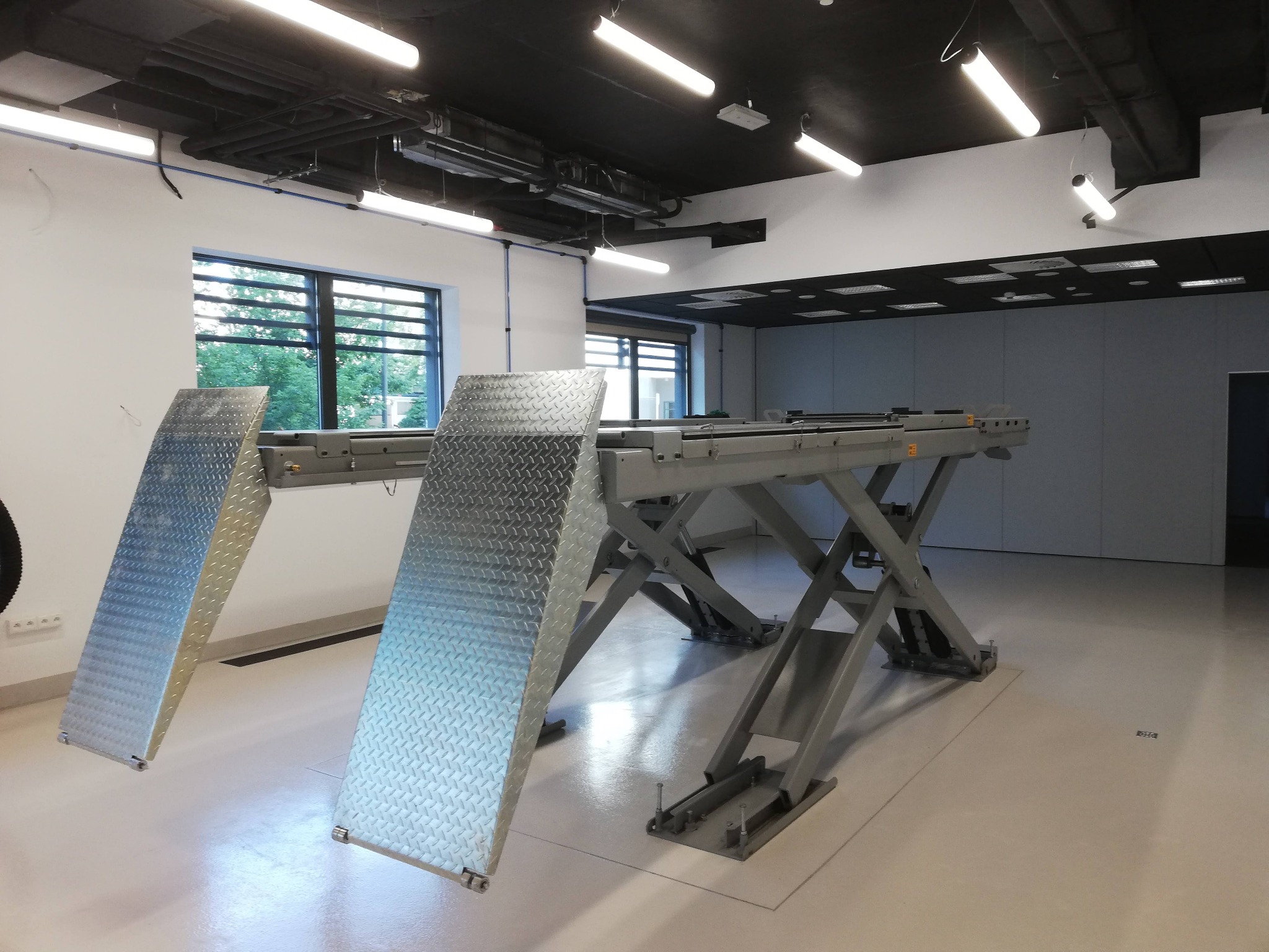 Warsztat samochodowy z podnośnikiem nożycowym w pozycji spoczynkowej, oświetlonymi lampami sufitowymi i oknem w tle.