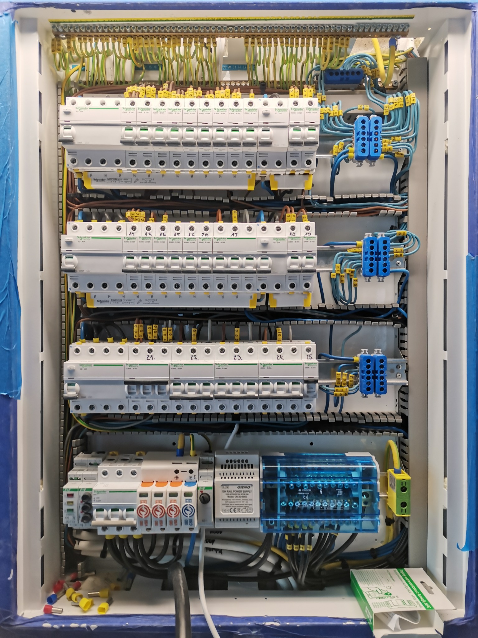 Rozdzielnica elektryczna z zabezpieczeniami i okablowaniem, widoczne liczne bezpieczniki, złączki i przewody w różnych kolorach, wewnątrz obudowy zabezpieczonej taśmą malarską.
